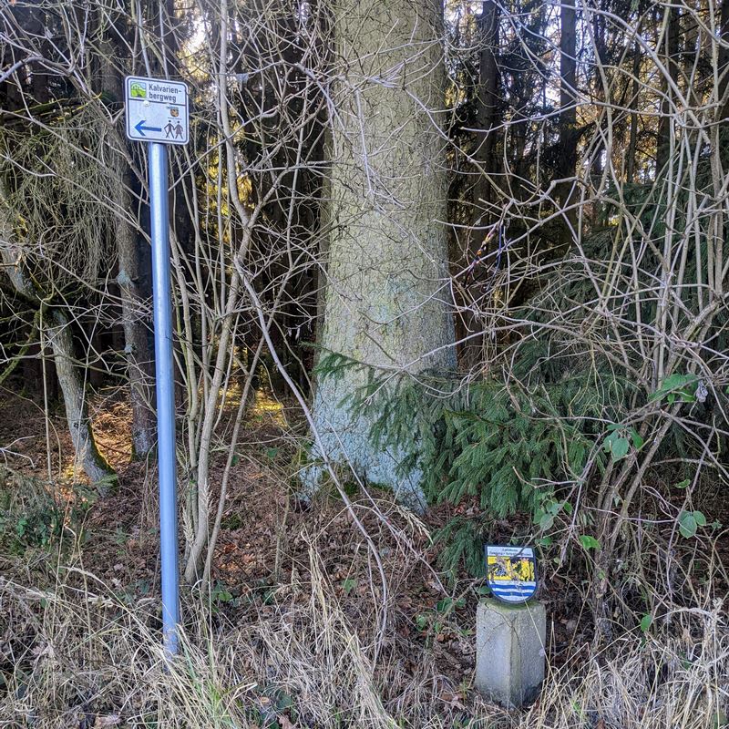 Grenzstein des Landkreises Neuburg- Schrobenhausen (rechts unten im Bild). Dahinter Wald bzw. Unterholz. AM linken Bildrad ein Wegweisen "Wanderweg Kalvarienberg".