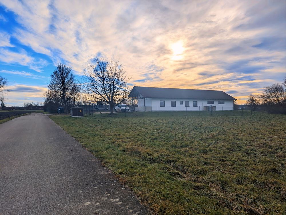 Die Ansicht der Kläranlage Karlskron aus nördlicher Richtung im winterlichen Licht der Nachmittagssonne. Das Bild zeigt ein fotorealistisches Bild eines weißen Scheunen-ähnlichen Gebäudes in ländlicher Umgebung. Das Gebäude hat ein Datteldach und ist von Feldern umgeben. Der Himmel ist blau mit weißen Wolken und die Sonne geht im Hintergrund unter. Das Bild wurde von einer Schotterstraße aufgenommen, die zum Gebäude führt. Im Hintergrund befinden sich Bäume und ein Zaun um das Gebäude.