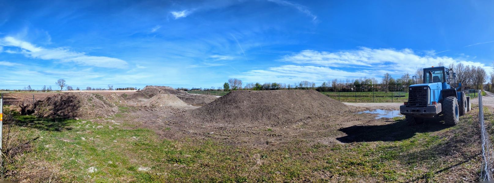 Auf dem Bild ist der Bikepark Pobenhausen im Bau zu sehen. Es handelt sich um eine Freizeiteinrichtung für Mountainbiker, die speziell für das Befahren mit Fahrrädern im Gelände konzipiert ist. Die Szene zeigt eine Baustelle mit Erdhügeln, Rampen und Strecken, die für zukünftige Mountainbike-Abenteuer gestaltet werden. Die Sonne scheint, und die Umgebung ist von grünen Bäumen und Wiesen umgeben.