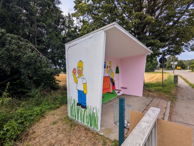 Das Bild zeigt eine Wandmalerei der animierten Fernsehserie “Die Simpsons”. Die Malerei befindet sich im Brautlacher Bushäuschen. Zu sehen ist die westliche Außenseitedes Bushäuschens. Homer betrachtet einen Donut, den er in der Hand hält. Im Hintergrund sind eine grasbewachsene Fläche, Bäume und ein offenes Feld zu sehen.