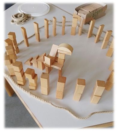 Eine aus Holzbausteinen gebaute Figur, die an einen Dinosaurier oder ein Fantasiewesen erinnert, steht in der Mitte eines Kreises aus aufrecht stehenden Dominosteinen und einem Seil auf einem hellen Tisch.